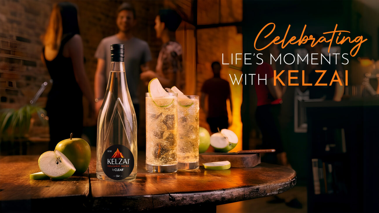 Kelzai volcanic water bottle on a wooden table with apple slices and sparkling drinks, set against a warm party background; premium sparkling water in India for celebrations.